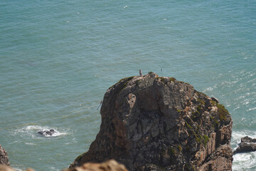 A rocky outcrop juts into the ocean. A few figures stand atop the rock, silhouetted against the bright sky and water. Waves crash against the base of the rock.