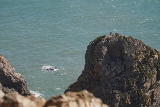 Two figures stand on a rocky cliff overlooking the turquoise ocean. Waves crash against the rocks below. The scene evokes a sense of solitude and vastness.