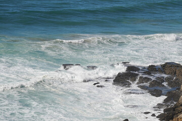 Waves crash against rocky shoreline. Turquoise water churns with white foam. Dark rocks are visible in the foreground. The scene evokes a sense of power and natural beauty.