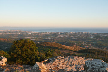 A scenic view from a hilltop shows a sprawling town and coastline bathed in the warm light of sunset. Lush greenery and rocky terrain fill the foreground.