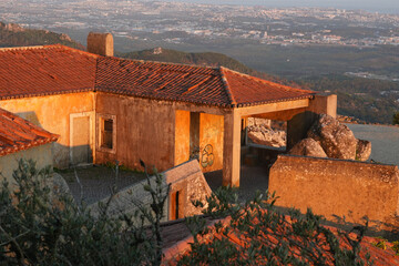 A rustic house with a terracotta roof sits atop a hill, overlooking a sprawling cityscape bathed in...