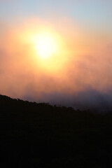 A dramatic, dark cliff face contrasts with a vibrant sunset. The sun shines brightly through a haze, creating a warm, orange glow against a pale sky.