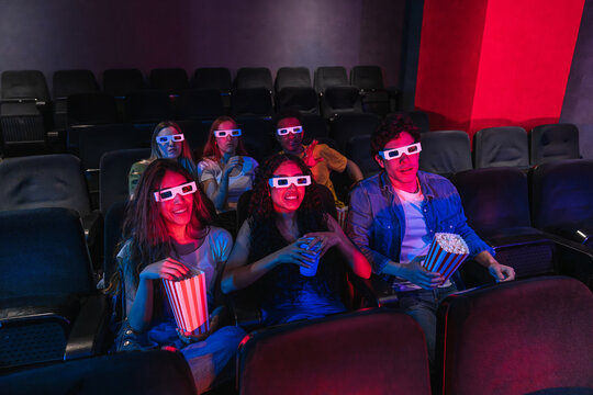 Group of young friends wearing 3d glasses, enjoying a film and munching on popcorn while relaxing in comfortable movie theater seats - Powered by Adobe