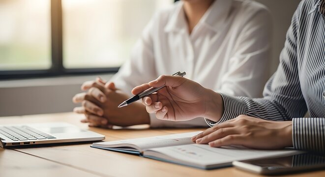 Two people in a meeting, one pointing at a document while the other listens attentively, with a laptop and phone on the table