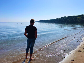 Slim man in jeans and top and bare feet on sandy Studland beach looking out to Old Harry Rock in Dorset UK.
