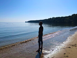 Slim man in jeans and top and bare feet on sandy Studland beach looking out to calm glassy sea.