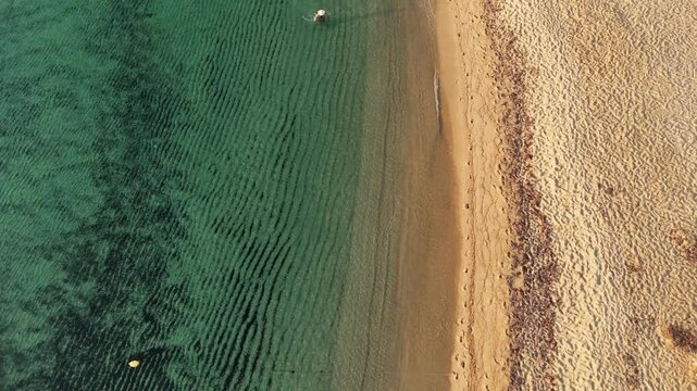 Stunning aerial drone view of a serene beachscape featuring crystal-clear greenish-blue waters gently meeting golden sand. The shoreline forms elegant wave patterns, while neatly arranged white beach 