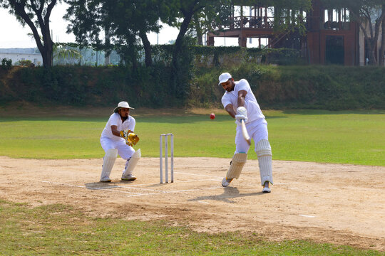 A cricketer in white hits a powerful shot, with a wicketkeeper ready behind the stumps on a sunny cricket field
