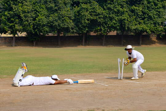 A cricketer in white dives to reach the crease, while a wicketkeeper prepares to catch the ball on a cricket field