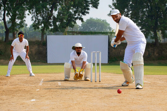 A batsman in white prepares to hit the ball, with a wicketkeeper and fielder ready, on a cricket field