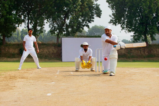 A batsman in white plays a shot as a wicketkeeper and fielder observe, on a bright cricket field
