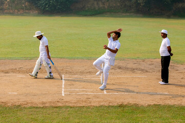 A bowler in white delivers a ball as a batsman and umpire watch, on a cricket field