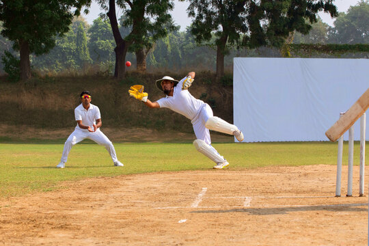 A wicketkeeper in white dives to catch the ball as a fielder watches, on a cricket field