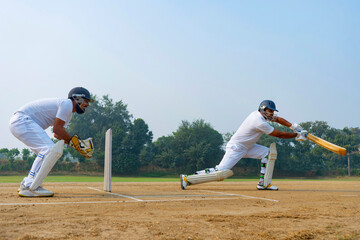 A batsman in white executes a powerful shot on a dirt field, with a wicketkeeper closely observing, highlighting teamwork and skill