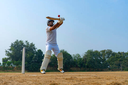 A cricketer in white aims for a high shot on a dirt field, showcasing power and precision under a clear sky