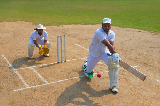 A batsman in white uniform executes a powerful shot, with a wicketkeeper crouched behind on a cricket field