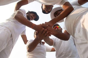 A cricket team in white huddles together, hands stacked, showing unity and teamwork, captured from a low angle