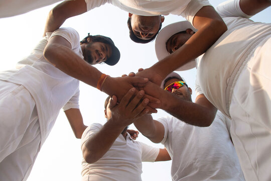 A cricket team in white huddles together, hands stacked, showing unity and teamwork, captured from a low angle