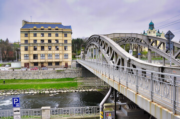 Ostrawa - Most Miloše Sýkory. Stalowy most łukowy przecina spokojną rzekę, obok beżowego miejskiego budynku i trawiastych brzegów. © Pol Badger