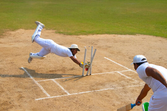 A wicketkeeper in white dives dramatically to break the stumps as a batsman watches, set on a cricket field under clear skies