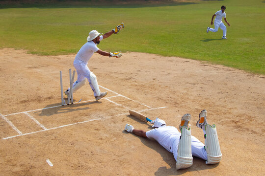 A cricketer in white appeals for a run-out as a batsman lies on the ground, with another player running in the background on a sunny day