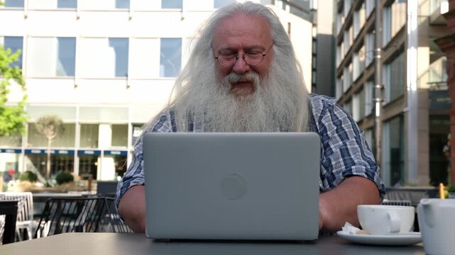Elderly man enjoys coffee while working on laptop outdoors in a lively city plaza during a sunny day