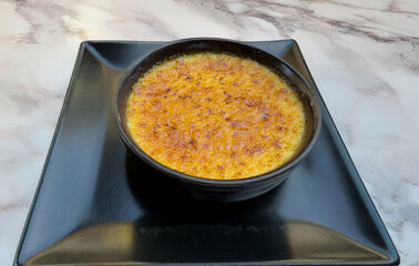 Caramelized Catalan cream dessert in black ceramic bowl on square plate