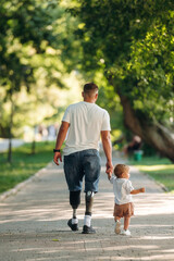 Rear view, from the back. Father that is amputee with prosthesis is with his little son in the park