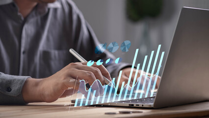 Close-up of businessman analyzing virtual financial charts and graphs on laptop with stylus pen, symbolizing business analytics, data visualization, investment planning, and economic growth concept.