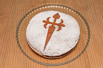 A modern version of Tarta de Santiago with a touch of cinnamon and brandy, maintaining the traditional cross design in contrast with the powdered sugar