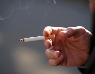 Close-up of a hand holding a lit cigarette with smoke rising against a blurred background
