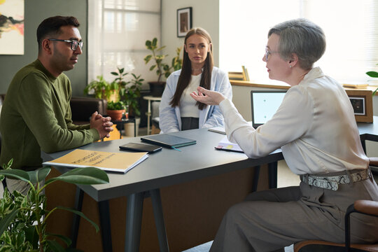 Middle aged woman psychologist conducting therapy session with South Asian man while woman observing in modern office setting