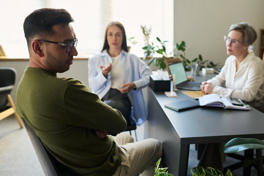 Man sitting with arms crossed facing away from two middle aged women, one gesturing while other taking notes during psychological counseling session