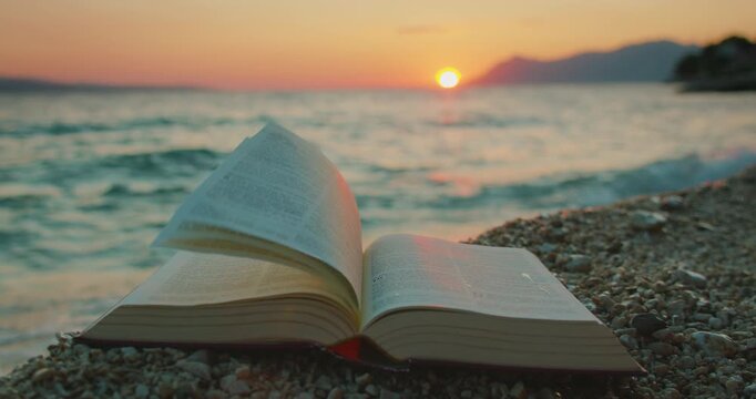 Open book Holy Bible rests on shore at sunset with sea and mountains in background