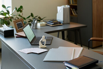 Modern psychologist office showing open laptop, abstract human figure sculpture, paperwork and folders on desk, background featuring organized workspace with plants and office supplies