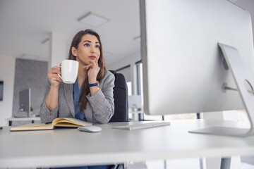 Woman sipping coffee while thinking at her workspace during a busy day in the modern office
