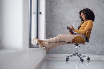 Woman sitting by window writing on tablet during afternoon in modern office space