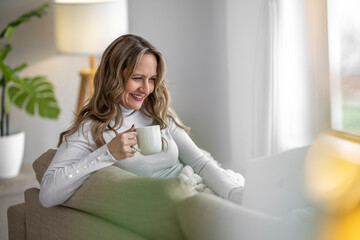 Woman enjoying a warm drink while using a laptop in a cozy living room during a bright afternoon