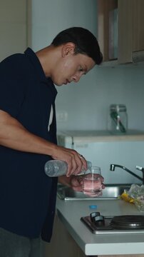 Young man standing near kitchen counter filling glass with fresh water from bottle and quenching thirst