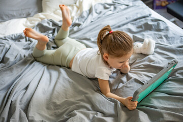 Little girl lying on her parents bed and watching cartoons on a tablet. Concept of screen time, digital entertainment and relaxation during early childhood.