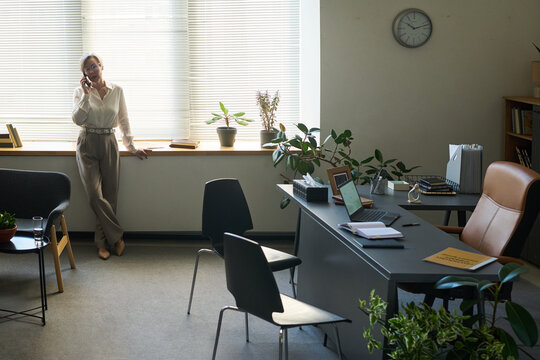 Middle aged woman standing by window talking on smartphone in psychologist office, desk with laptop and documents in foreground, plants and chairs visible in workspace