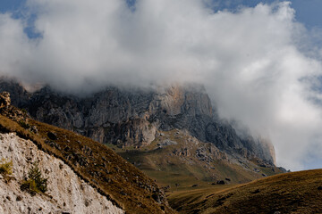 A rugged mountain landscape with steep cliffs and a cloudy sky. The terrain is rocky with patches of grass and shrubs.