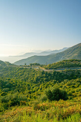 Fototapeta premium Scenic hillside with winding road leading towards sea and layered mountain ridges fading in horizon under bright blue sky in warm daylight