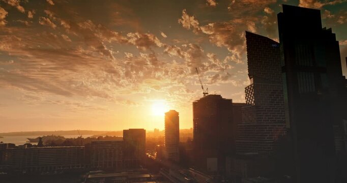 Australia, Sydney: Sydney skyscrapers and harbor at sunset. Warm orange sunlight illuminates buildings, creating stunning cityscape silhouette against backdrop of dramatic clouds. Drone flight footage