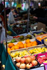 Fresh pomegranates, oranges, and apples are for sale in wooden crates at a street fruit market in Paris, France. A sign for a fruit brand is in the foreground.