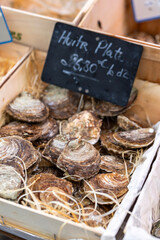 Fresh oysters of the Huitr Plate variety are for sale in a wooden crate at a street seafood market in Paris, France. A chalkboard sign with the name and price is in the background.