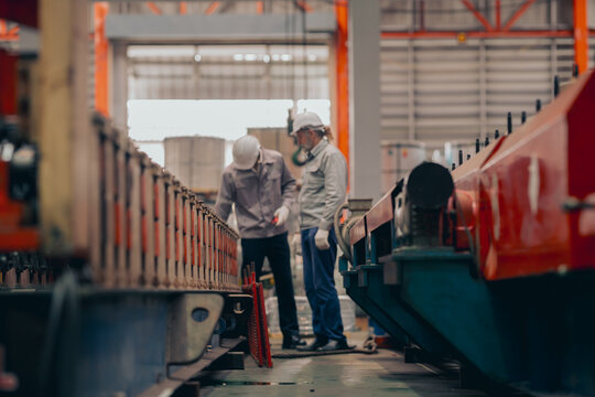 Professional mature engineer in safety hardhat and vest inspecting metal parts and machinery operations in a large industrial manufacturing facility. Highlighting technical expertise, safety standards