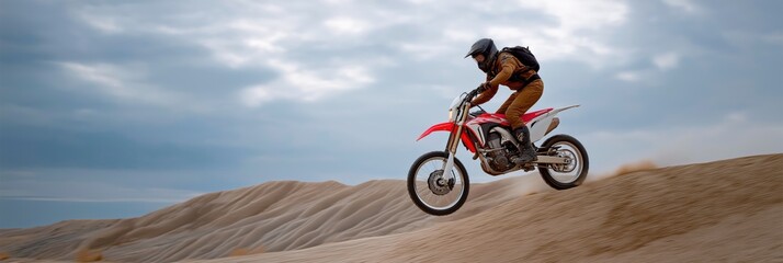 Young hispanic male motorcyclist jumping dirt bike on sand dunes under cloudy sky