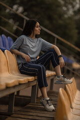 brunette girl relaxes alone in the empty stands of a stadium. She wears a blue t shirt and casual pants enjoying a peaceful moment in the sports venue.