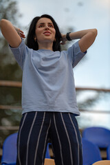 A beautiful brunette girl stands in an empty stadium smiling widely while raising her arms with excitement. It is a clear day and she feels triumphant.